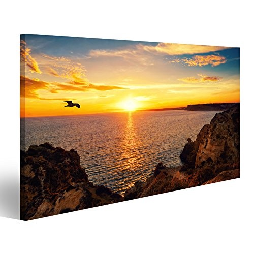 Bild auf Leinwand Ruhige Sonnenuntergangskulisse am Meer mit dem auf dem Wasser reflektierten Sonnenlicht, Einem fliegenden Vogel und der felsigen Küste. Wandbild Poster Leinwandbild