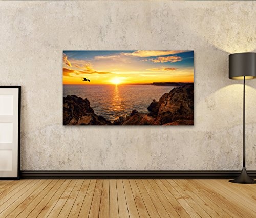 Bild auf Leinwand Ruhige Sonnenuntergangskulisse am Meer mit dem auf dem Wasser reflektierten Sonnenlicht, Einem fliegenden Vogel und der felsigen Küste. Wandbild Poster Leinwandbild